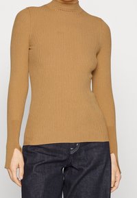 Ribbed turtleneck-kardigan vaaleanruskeana, valmistettu pehmeästä neulostyöstä. Ominaisuuksina pitkät hihat, joissa on halkiot, ja kapea mitoitus.