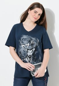 Námořnická modrá V-neck tričko s krátkým rukávem s grafickým vyobrazením jelena a textem. Modelka drží stříbrnou metalickou kabelku, čímž ukazuje neformální styl.