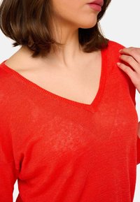 Femme aux cheveux courts bruns portant un pull V rouge vif, ajustant la zone des épaules avec la main droite.