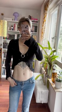 Zwarte langemouwen top met een diepe V-hals en hartvormige accent, gecombineerd met lichtblauwe jeans. Binnenomgeving met planten en boekenplanken.