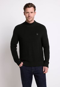 Pull en maille noire avec un col montant et une texture côtelée. Doté d'un petit logo circulaire sur la poitrine. Associé à un jean en denim sombre.
