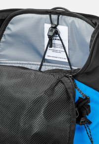 Svart nätfack inuti en ryggsäck, med grå foder, en vit etikett med svart text och blå detaljer synliga på utsidan.