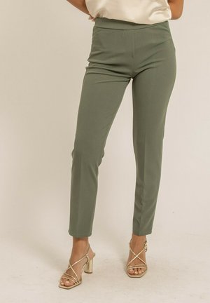 Pantalon tailleur vert avec une texture lisse et une coupe ajustée, doté d'une taille haute. Associé à des talons dorés à lanières sur un fond neutre.