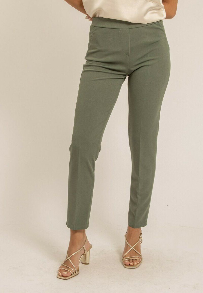 Pantalon tailleur vert avec une texture lisse et une coupe ajustée, doté d'une taille haute. Associé à des talons dorés à lanières sur un fond neutre.