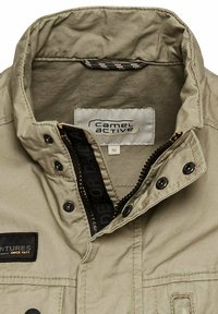 Beige Jacke mit hohem Kragen, Reißverschluss vorne und Druckknöpfen. Verfügt über ein schwarzes Etikett mit "Camel Active" und strukturiertem Stoff.
