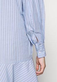 Chemise bleu clair à manches longues avec fines rayures noires verticales, poignet boutonné, et une main détendue le long du corps sur un fond uni.