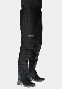 Zwarte geïsoleerde broek met een doorgestikt ontwerp, ritszakken en een elastische taille. Kenmerkt zich door een logo op de linkeronderkant van het been. Draagt zwart schoeisel.