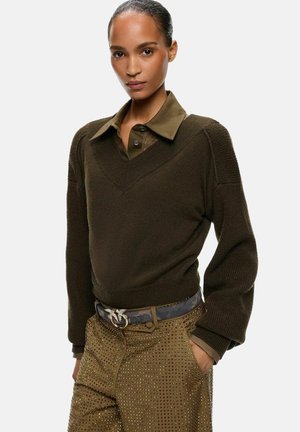 Donna con la pelle scura e capelli tirati indietro, che indossa un maglione verde scuro sopra una camicia con colletto e pantaloni marroni borchiati con cintura.