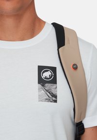 T-shirt bianco a maniche corte con una patch grafica nera raffigurante un mammut e un motivo a linee ondulate; indossato sotto una spallina beige.