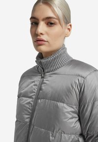 Doudoune grise avec un col montant côtelé, dotée d'une fermeture éclair, d'un motif matelassé et d'une texture lisse et brillante.