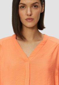 Blouse en V couleur corail vif, fabriquée dans un tissu léger et froissé. Manches courtes et détail subtil de patte de boutonnage à l'avant.
