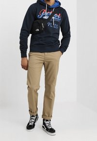 Mand iført navy hættetrøje med logo, beige bukser med opbukket kant, sorte høje sneakers og sort skuldertaske mod en ensfarvet hvid baggrund.