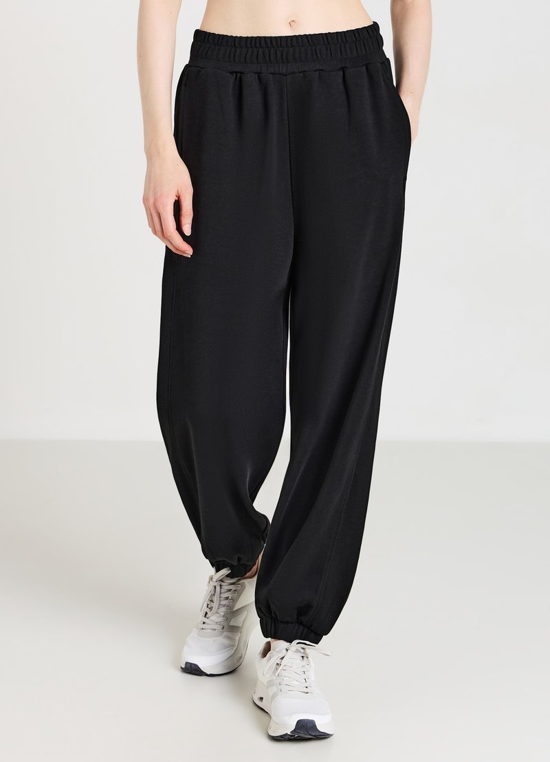 Pantalons de jogging noirs amples avec une taille élastique et des poignets élastiques. Le tissu semble lisse avec un léger éclat. Portés avec des baskets blanches.