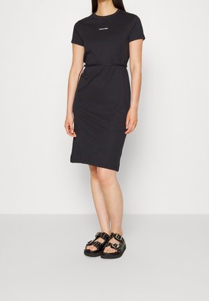 Woman wearing a black Calvin Klein knee-length dress with a thin belt and black studded buckle sandals standing against a white background.