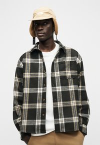 Νέος άνδρας που φοράει μπεζ καπέλο με αυτιά, πουκάμισο καρό σε μαύρο και μπεζ πάνω από λευκό T-shirt και καφέ παντελόνι, σε απλό φόντο.