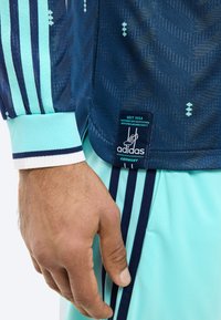 Gros plan sur la main d'une personne portant un maillot de sport Adidas bleu et bleu clair ainsi que des shorts avec le logo de l'équipe d'Allemagne et trois bandes bleu marine.