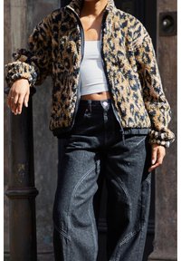 Personne portant une veste en polaire imprimé léopard par-dessus un crop top blanc et un jean noir taille haute, s'appuyant sur un poteau en métal à l'extérieur.