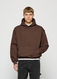 Junger Mann in einem braunen Kapuzenpullover und schwarzen Jeans, mit der rechten Hand in der vorderen Tasche, stehend vor einem schlichten grauen Hintergrund.