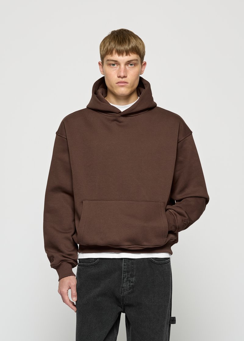 Junger Mann in einem braunen Kapuzenpullover und schwarzen Jeans, mit der rechten Hand in der vorderen Tasche, stehend vor einem schlichten grauen Hintergrund.