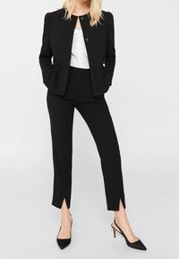 Vrouw in zwarte op maat gemaakte cropped broek met voorflitsen, zwarte gestructureerde blazer, witte blouse en zwarte slingback hakken, staande met haar handen in de zakken.