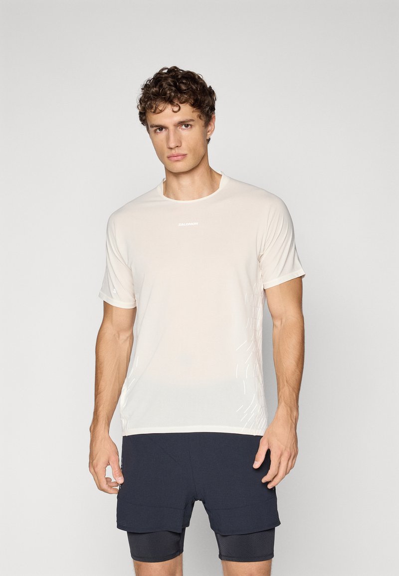 Man with curly hair wearing a light beige athletic t-shirt and layered dark shorts, standing against a plain light background.