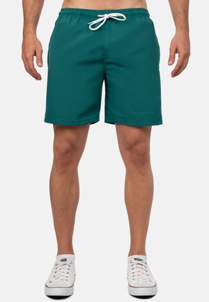 Mężczyzna w turkusowych spodenkach kąpielowych z białym sznurkiem i białych niskich tenisówkach stoi na jednolitym tle.