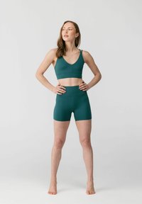 Jalgrattasõiduks sobivas tumerohelises spordirinnahoidjas ja sobivate kõrge vöökohaga lühikeste pükstega paljajalu seisvad naine, käed puusal, ühetoonilise heleda tausta ees.