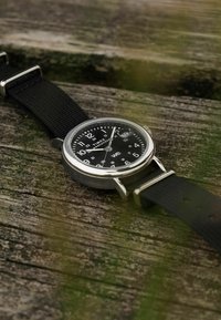 Timex Weekender kellaga, millel on must tahvel ja kangast rihm, puhkamas kulunud puidust pinnal, aeg umbes 1:57.