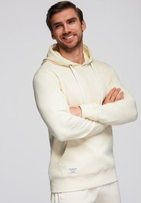 Homme souriant, bras croisés, portant un sweat à capuche crème et un pantalon assorti, sur un fond gris uni.