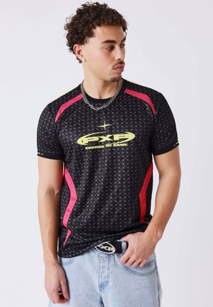 Hombre con cabello rizado que lleva una camiseta deportiva negra y roja con el logo amarillo "FXP Change The Game" y vaqueros azul claro, mirando hacia un lado.