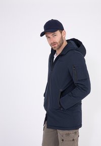 Marineblaues Kapuzenjacke mit Reißverschluss, Seitentaschen und einer Reißverschlusstasche an den Ärmeln, aus einem glatten, matten Stoff gefertigt.