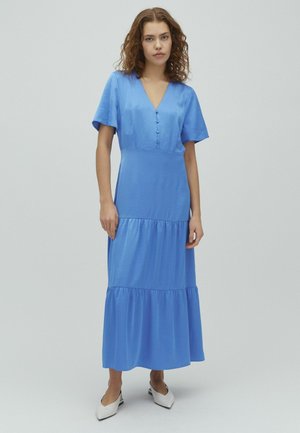 Göndör hajú nő, kék, gombos, V-nyakú, réteges midi ruha és fehér, hegyes orrú slingback cipő viselése mellett, egyszerű háttér előtt áll.