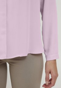 Blusa a maniche lunghe rosa chiaro realizzata in tessuto morbido, con orlo dritto, pattina con bottoni e maniche con polsini, abbinata a pantaloni beige.