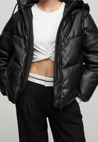 Chaqueta puffer negra con un acabado brillante, capucha y cierre de cremallera. Lleva puesta una camiseta blanca corta, combinada con pantalones negros de cintura alta.