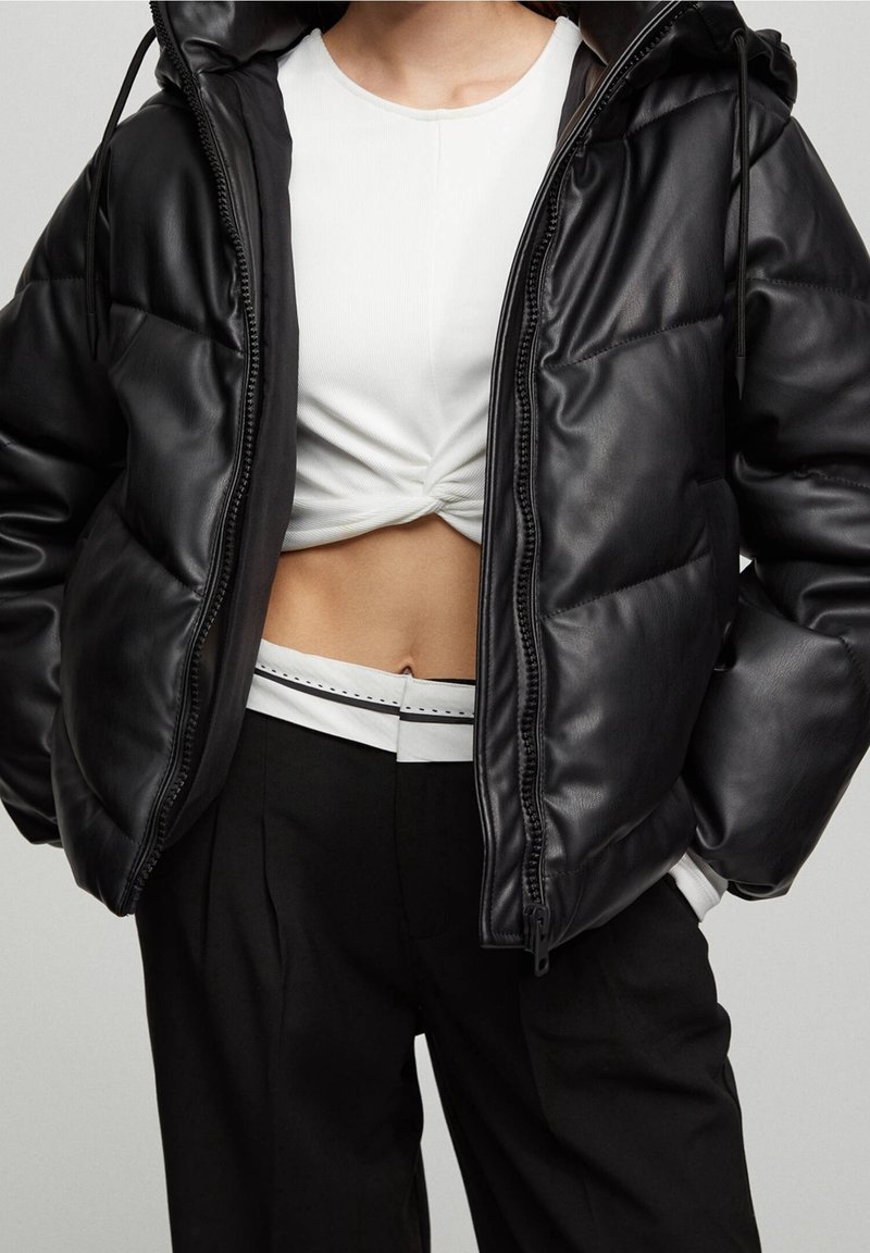 Chaqueta puffer negra con un acabado brillante, capucha y cierre de cremallera. Lleva puesta una camiseta blanca corta, combinada con pantalones negros de cintura alta.