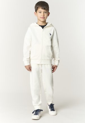 Ragazzo in piedi, che indossa una felpa bianca con zip e piccolo logo, pantaloni della tuta bianchi abbinati e sneakers bianche con lacci scuri, sfondo neutro.