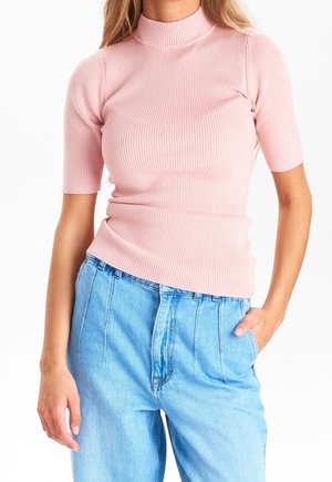 Femme portant un top à col roulé côtelé rose ajusté à manches courtes et un jean bleu clair taille haute, une main dans la poche.