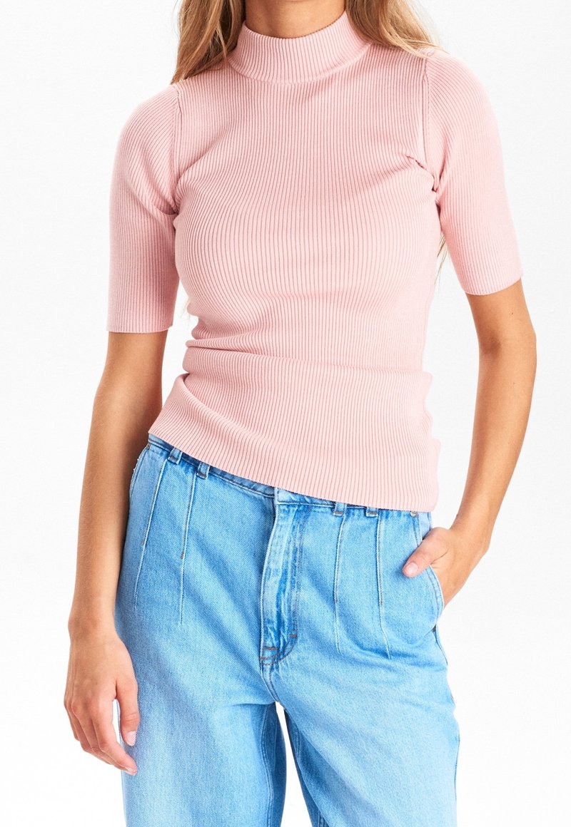 Femme portant un top à col roulé côtelé rose ajusté à manches courtes et un jean bleu clair taille haute, une main dans la poche.