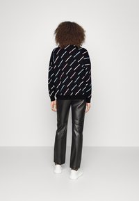 Una persona con capelli ricci sta in piedi di spalle, indossando una felpa nera con testo colorato diagonale e pantaloni di pelle nera con scarpe da ginnastica bianche.