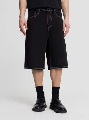 Uomo che indossa pantaloncini neri fino al ginocchio con cuciture bianche, calzini neri e mocassini neri lucidi, in piedi contro uno sfondo semplice.