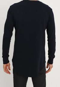 Námořnicky modrá košile s dlouhým rukávem a kulatým výstřihem, se zakřiveným spodním lemem a měkkým, hladkým materiálem. Viditelné švy na manžetách a lemu.