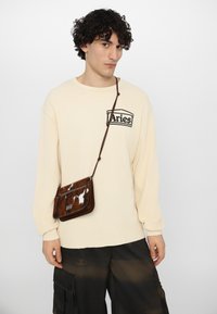 Sac bandoulière en cuir de vache marron et crème avec une finition texturée, doté d'une fermeture à rabat et d'une sangle réglable ; porté sur une chemise à manches longues crème.