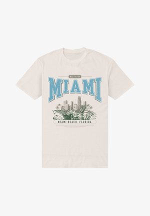 T-shirt en coton crème à manches courtes avec un graphique bleu indiquant "PROCHAINE STOP MIAMI" et des illustrations de skyline urbain et de plantes tropicales.