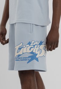 Světle modré bavlněné šortky mají velký bílý a modrý grafický potisk se stylizovaným textem a hvězdnými akcenty, včetně značkového loga.