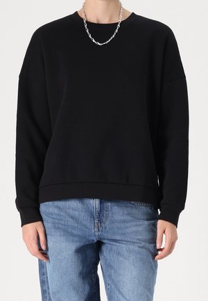 Osoba ubrana w gładką czarną bluzę z długim rękawem, jasnoniebieskie dżinsy oraz srebrny łańcuszek, stojąca na jednolitym tle.