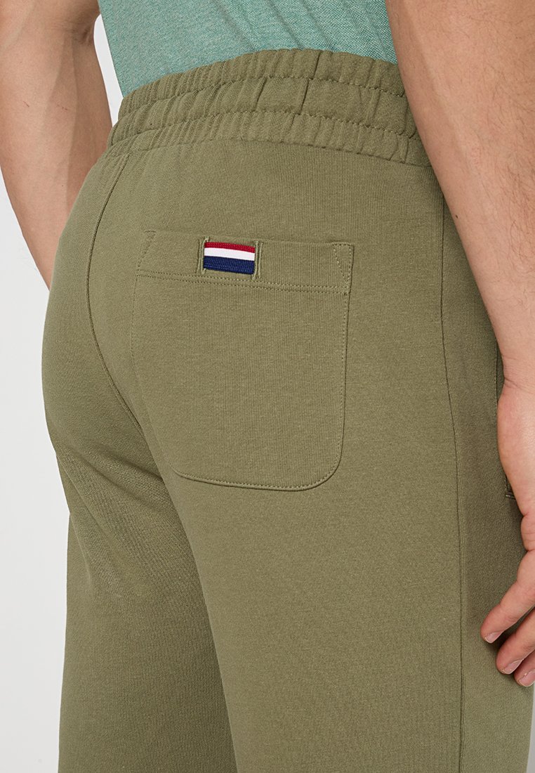 Olivgrüne Jogginghose mit elastischem Bund, ausgestattet mit einer Gesäßtasche und einem kleinen rot-weißen-blauen Akzentstreifen auf der Tasche.