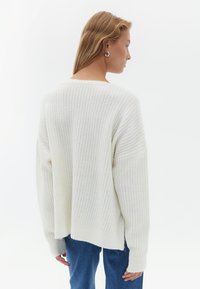 Crème gebreide trui met verticale ribbels, verlaagde schouders en brede mouwen. Opvallend door de ruime pasvorm en zachte textuur. Gepaard met blauwe jeans.