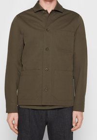 Giacca verde oliva in cotone con colletto, bottoni frontali e due tasche a toppa. Presenta una texture liscia e una vestibilità su misura.
