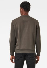 Sudadera de color verde oliva con cuello redondo, puños y dobladillo acanalados. Textura suave, con un parche rectangular en la parte superior de la espalda.