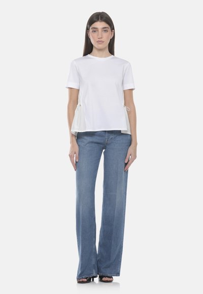Femme portant un haut blanc à manches courtes avec des cordons latéraux et un jean bleu à jambes larges, debout devant un fond uni.